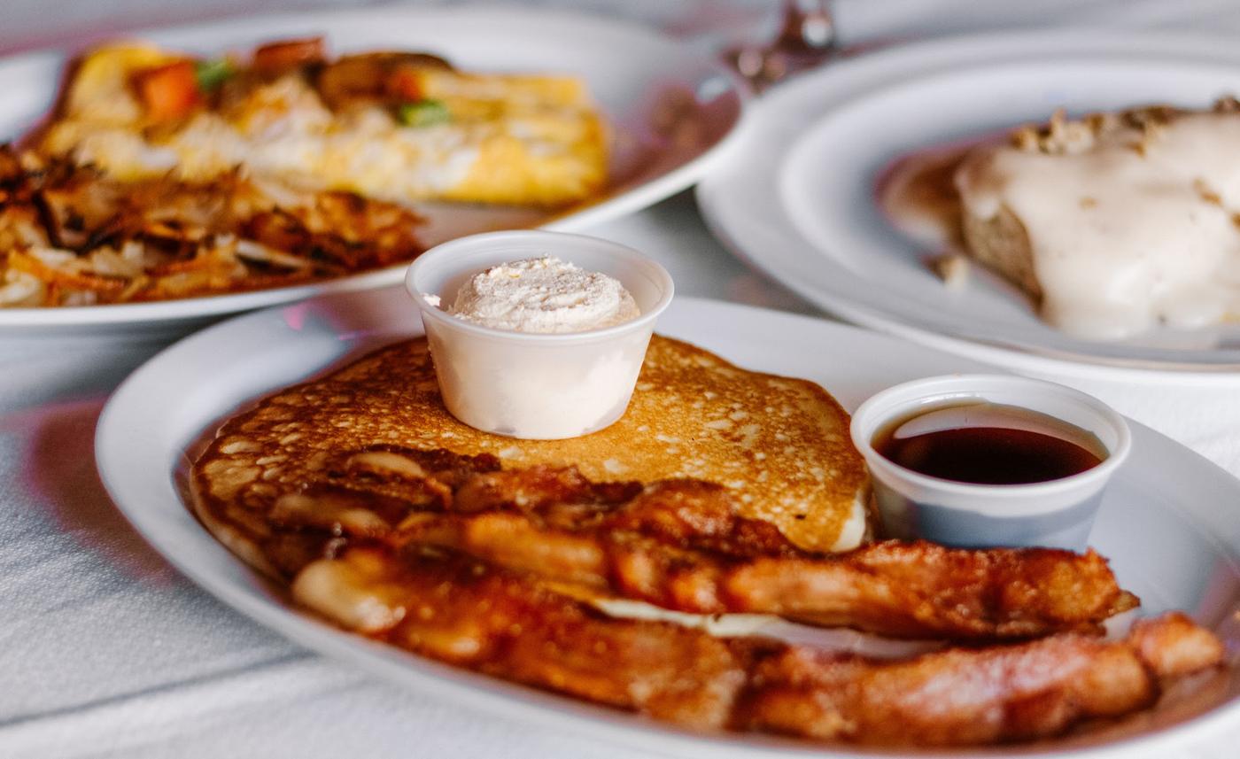 Pancakes with bacon, whipped butter and syrup, with an omelet and hash browns behind