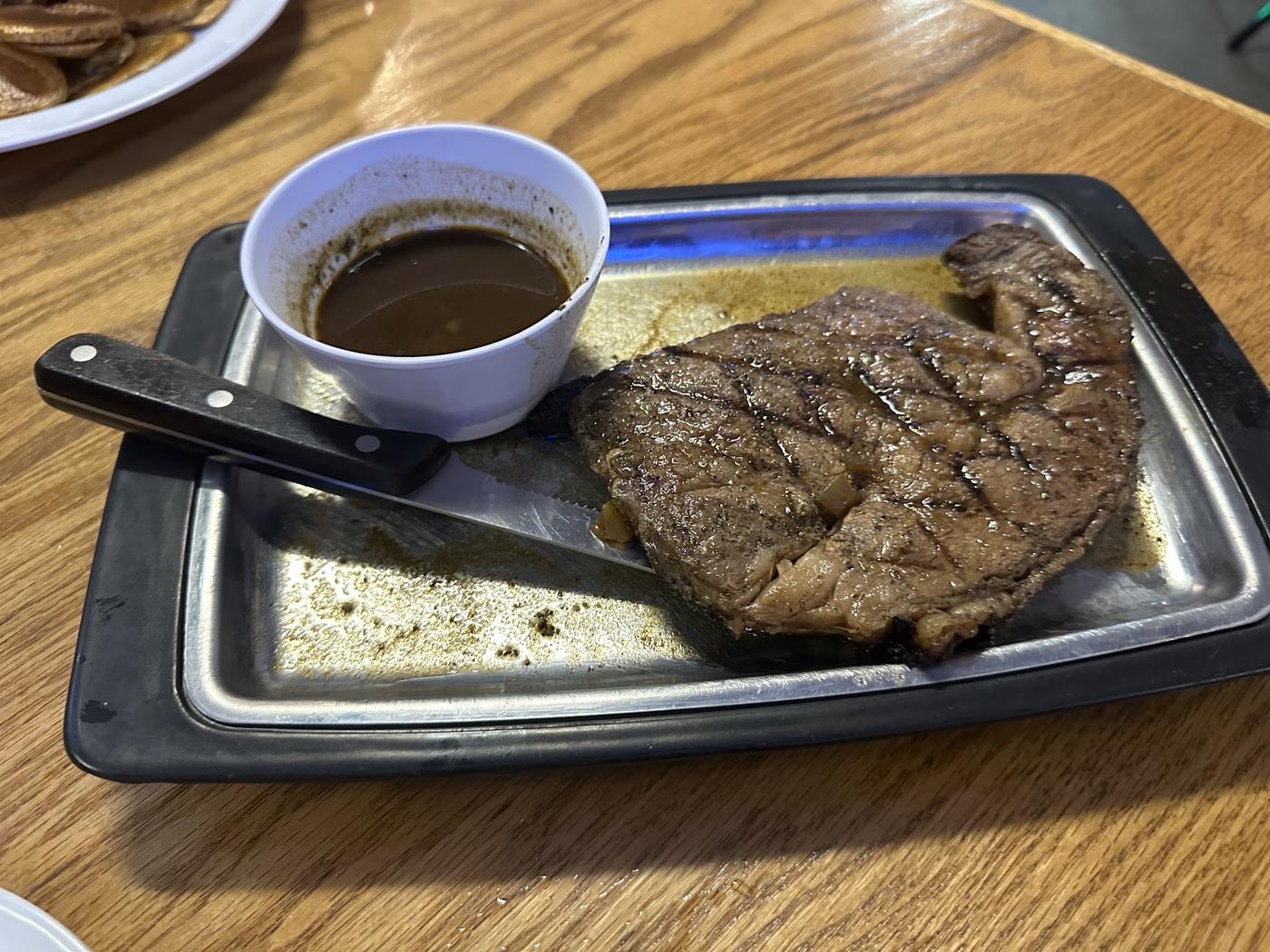A steak on a metal sizzle platter with a cup of au jus, on a wooden table